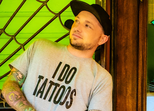 Travis wearing a gray t-shirt with the words 'I DO TATTOOS.' Travis is looking off to the side with a slight smile, giving off a relaxed and approachable vibe.