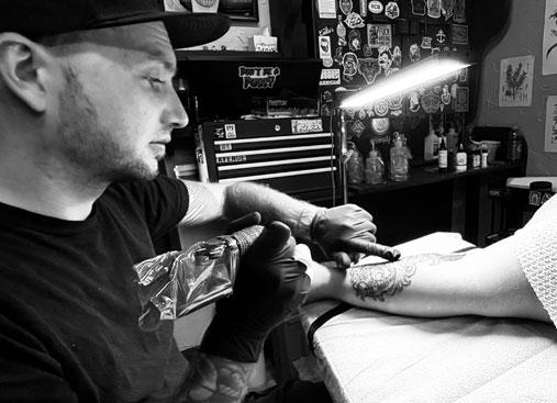 A black and white image showing Travis the tattoo artist working on the arm of a client.