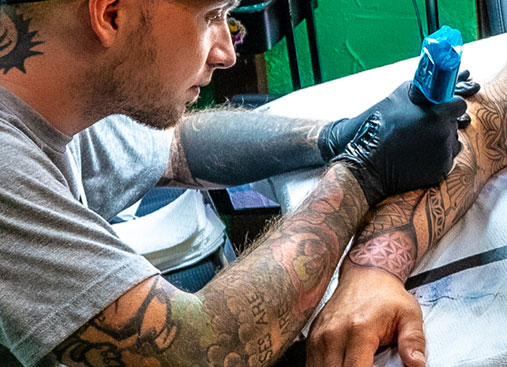 Travis carefully applies ink to a client's arm. Travis is working on a detailed, geometric design with serious concentration, professionalism and focused, reflecting the precise and delicate work being performed.