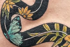 The tattoo depicts a stylized black snake adorned with various natural elements like flowers, a frog, a butterfly, and ladybugs.