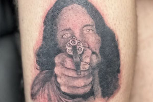 The tattoo depicts a person holding a gun, pointing it directly at the viewer.